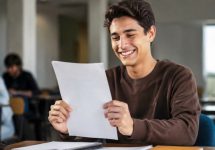 Um jovem estudante sorridente segura sua prova com expressão de felicidade e alívio após ser aprovado, em uma sala de aula iluminada naturalmente, com outros alunos ao fundo