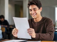 Um jovem estudante sorridente segura sua prova com expressão de felicidade e alívio após ser aprovado, em uma sala de aula iluminada naturalmente, com outros alunos ao fundo