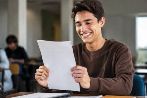 Um jovem estudante sorridente segura sua prova com expressão de felicidade e alívio após ser aprovado, em uma sala de aula iluminada naturalmente, com outros alunos ao fundo