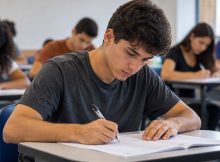 Jovem concentrado realizando uma prova em sala de aula, com outros estudantes ao fundo, em um ambiente realista e bem iluminado. Você gosta desta personalidade?